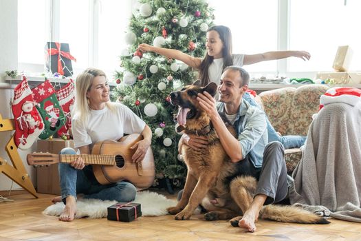 Friendly family is playing with dog near Christmas tree. They are sitting and laughing.