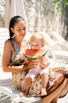 Little girl eats a piece of watermelon while sitting in her mother arms. High quality photo