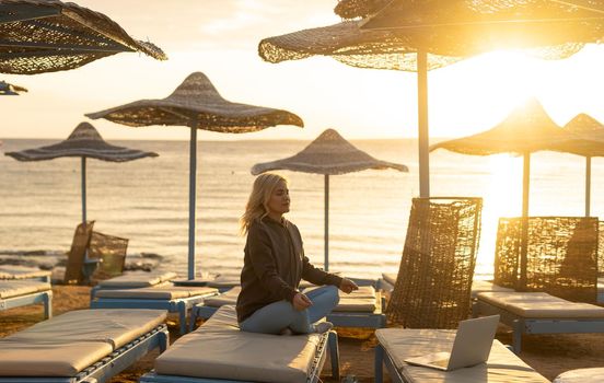 A calm woman sits on a sun lounger near the sea, gets distracted from working at the computer, meditates and removes negative emotions, combines remote work with rest