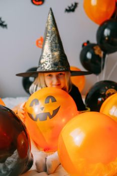 Children's Halloween - a girl in a witch hat and a carnival costume with airy orange and black balloons at home. Ready to celebrate Halloween.