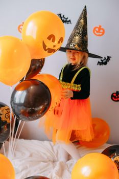 Children's Halloween - a girl in a witch hat and a carnival costume with airy orange and black balloons at home. Ready to celebrate Halloween.