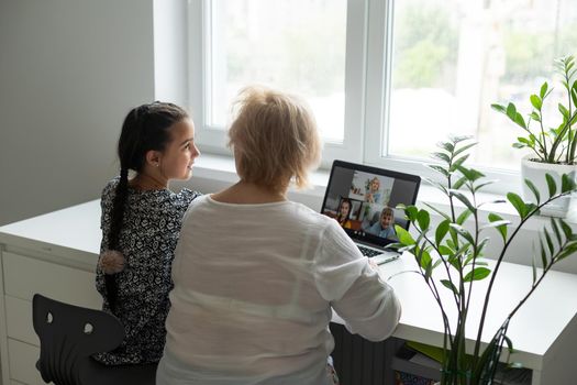 Portrait of happy grandmother and little granddaughter making video conference on pc sitting at table, waving hands at screen, greeting somebody, chatting with parents, enjoying online communication