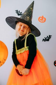 Children's Halloween - a girl in a witch hat and a carnival costume with airy orange and black balloons at home. Ready to celebrate Halloween.