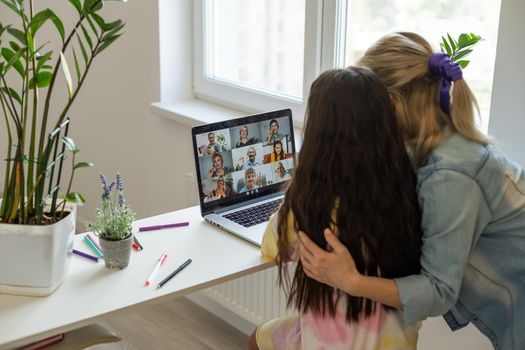 Cheerful little schoolgirl talking to the teacher while studying remotely via laptop at home with happy mom sitting nearby and giving support.