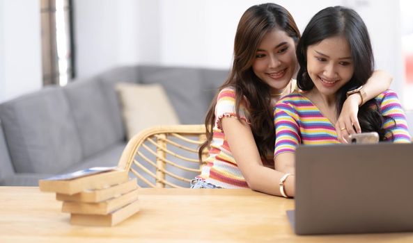 Young beautiful Asian women lesbian couple lover using smartphone and laptop shopping online together in living room at home with smiling face.Concept of LGBT sexuality with happy lifestyle together..