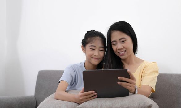 Happy young asian mom and daughter using digital tablet, watching videos or surfing internet, sitting on sofa at home