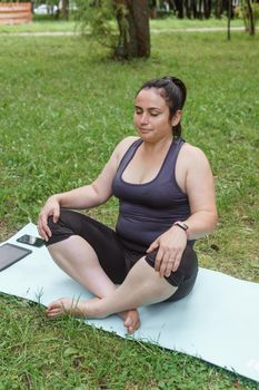 A charming brunette woman plus-size body positive practices sports in nature. Woman does yoga and meditation in the park on a sports mat