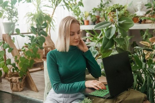 A blonde woman in a room with a lot of green indoor plants is working on a laptop. The concept of biophysical design in the interior. Work from home, work as a freelancer.