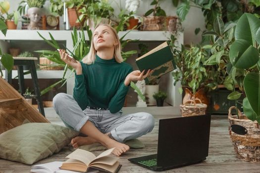 A blonde woman in a room with a lot of green indoor plants is working on a laptop. The concept of biophysical design in the interior. Work from home, work as a freelancer.