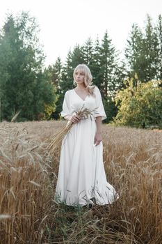A blonde woman in a long white dress walks in a wheat field. The concept of a wedding and walking in nature.
