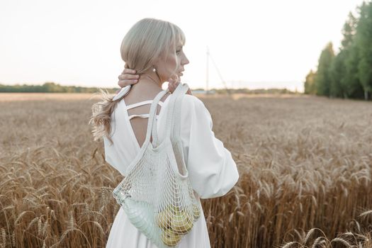 A blonde woman in a long white dress walks in a wheat field. The concept of a wedding and walking in nature.