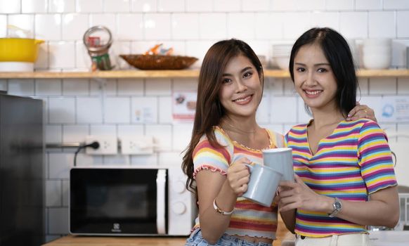 lesbian couple holding coffee mugs in kitchen looking at camera.