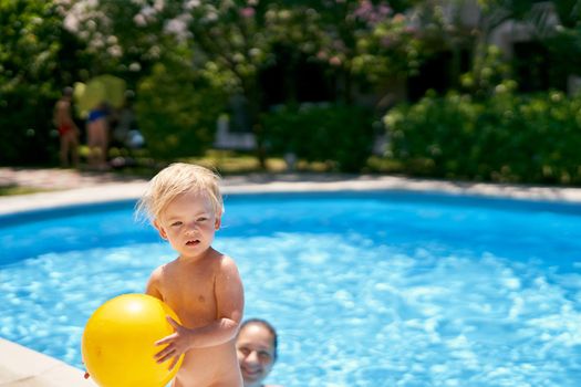 Small baby with a yellow ball stands by the pool with turquoise water. High quality photo