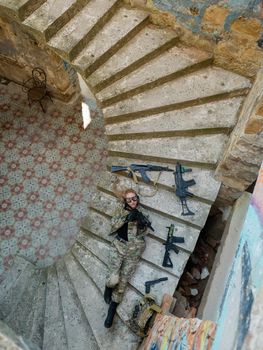 Caucasian woman in military uniform lies on the stairs of an abandoned building and holds a machine gun. View from above