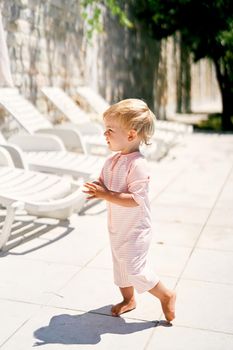 Little girl in summer overalls stands on a tile near white sun loungers. High quality photo