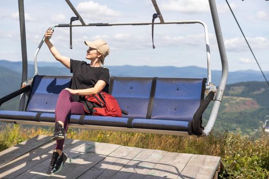 Young women sit on the Chairlift. They take pictures with smartphones. View from the back. Green grass and trees. Concept of a summer vacation in the mountains