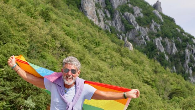 Portrait of a gray-haired senior elderly Caucasian man bisexuality with a beard and sunglasses holding a rainbow LGBTQIA flag on nature. Celebrates Pride Month, Rainbow Flag Day, gay parade