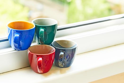 a small bowl shaped container for drinking from, typically having a handle.Four multi colored ceramic cups on the windowsill.Coffee and tea relaxation time