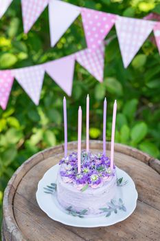 Very beautiful bento cake with purple, veri peri, matthiola flowers with green leaves, candles in the cake on the background of pink flags. Birthday. Vertical photo