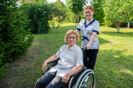 Caucasian female doctor walks with an elderly patient in a wheelchair in the park. Nurse accompanies an old woman on a walk outdoors