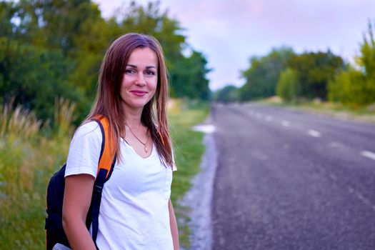 going or being transported from place to place.Young traveling cheerful girl stands waiting by the asphalt road with trees