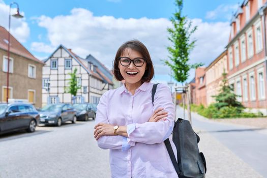 Portrait of confident successful mature woman on street of European city. Smiling beautiful middle aged female with crossed arms looking at camera