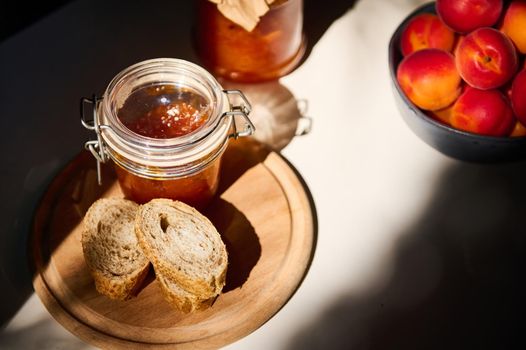 Still life, illuminated by a daylight. Top view. Slices of whole grain bread on wooden board, jar with homemade jam and ripe juicy apricots in blue ceramic bowl on white table in rustic summer kitchen