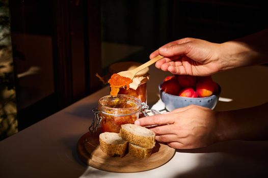 Food and drink, summer autumn harvest concept. Close-up of the hands of a housewife holding a wooden spoon and spreading homemade apricot jam on bread. Image illuminated by daylight.
