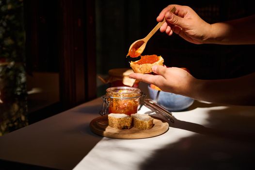 Sunlight illuminates the hands of a housewife spreading marmalade on bread in the summer village kitchen, while standing by the wooden window overlooking the garden. Close-up. Rural still life