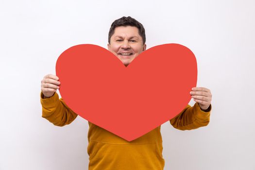 Handsome man holding big red heart, expressing positive romantic emotions, looking at camera with happy expression, wearing urban style hoodie. Indoor studio shot isolated on white background.