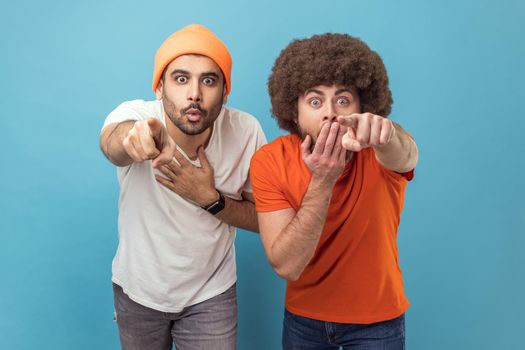 Wow, look at this. Portrait of two astonished surprised young adult hipster men looking at camera with big eyes, pointing at camera with fingers. Indoor studio shot isolated on blue background.