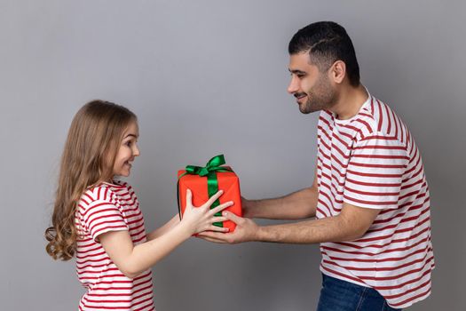 Portrait of satisfied father and daughter in striped T-shirts, man giving present box to his cute child, celebrating birthday together. Indoor studio shot isolated on gray background.