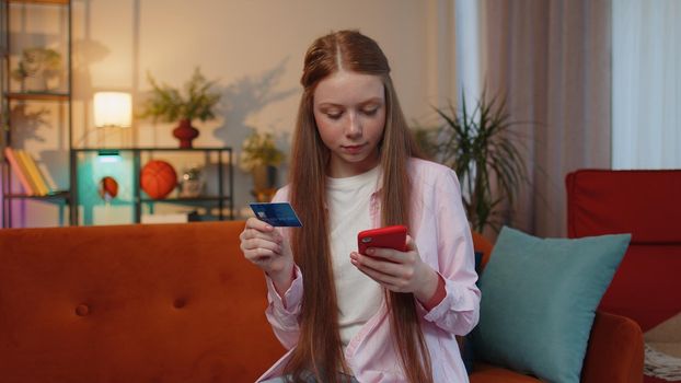 Happy shopaholic consumer young redhead teen child girl sitting at home making online order food delivery, payment with credit plastic bank card. Discount sale on internet. Ginger female children kid