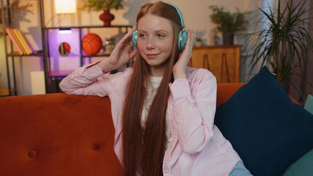 Happy relaxed overjoyed ginger kid in wireless headphones dancing on couch at home choosing listening favorite energetic disco rock n roll music in smartphone entertaining. People weekend activities