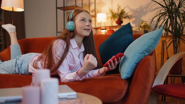 Happy excited joyful young redhead teenager child girl use mobile smartphone typing browsing say Wow yes found out great big win good news celebrate sitting at home couch. Ginger female children kid