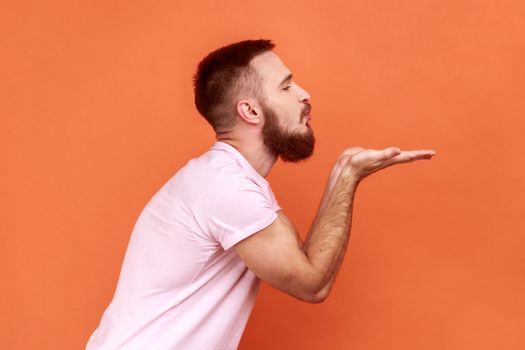 Side view portrait of handsome romantic young adult bearded man standing and looking ahead and sending air kiss, wearing pink T-shirt. Indoor studio shot isolated on orange background.