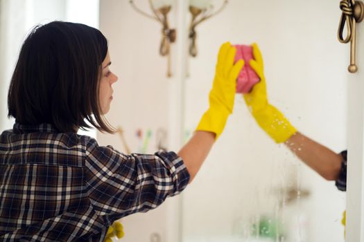 A young girl is cleaning the bathroom, applying detergent with a spray and washing the mirror with a sponge in yellow gloves on her hands. Woman taking care of the cleanliness of her home.