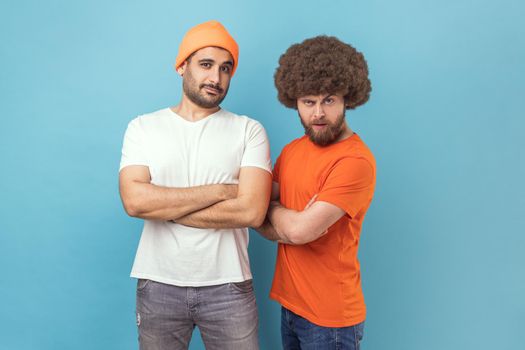 Portrait of two handsome serious young adult hipster men standing looking at camera with assertive expression, keeping hands folded. Indoor studio shot isolated on blue background.