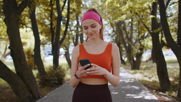 Athletic fitness sport runner girl sportswoman walking and using smartphone listening music with wireless earphones. Workout cardio in morning city park. Young woman jogger enjoying healthy lifestyle