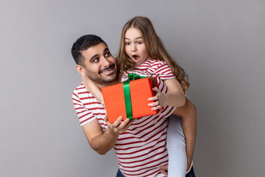 Portrait of pleased father holding his surprised daughter on his back and giving her a present for birthday, family wearing striped T-shirts. Indoor studio shot isolated on gray background.