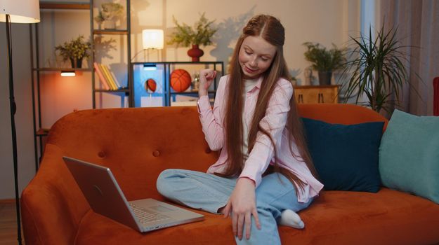 Overjoyed young child girl on home couch with laptop scream in delight raise hands in triumph winner celebrate success jackpot win money in lottery. Excited ginger children kid get online good news