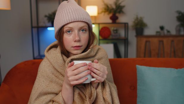 Sick child girl wear hat wrapped in plaid sit alone shivering from cold on sofa drinking hot tea in unheated apartment without heating due debt. Unhealthy children feeling discomfort try to warming up