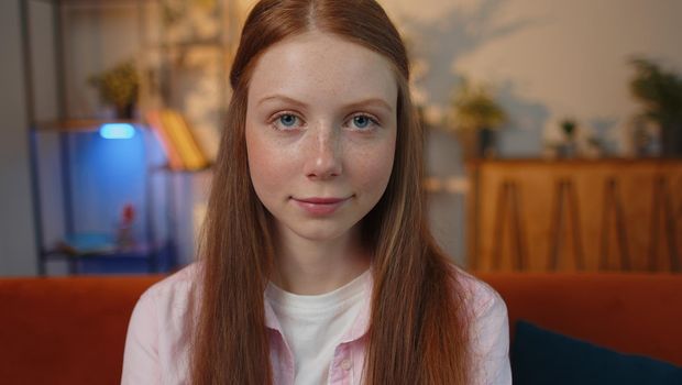 Close-up portrait of happy positive smiling young redhead child girl, looking at camera. Teen red-haired ginger freckles face kid indoor isolated at home in living room on couch. Female nature beauty