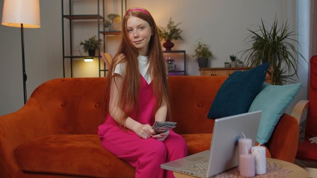 Planning budget. Rich happy redhead girl counting money cash use laptop computer calculate domestic bills at home. Joyful child satisfied of income earnings, saves money for planned vacation, gifts