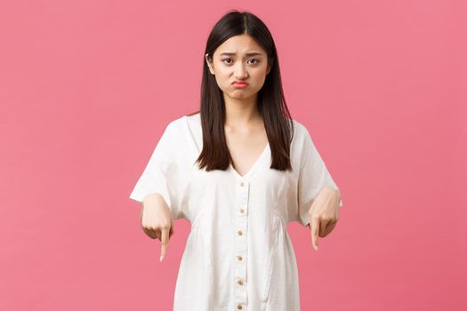 Beauty, people emotions and summer leisure and vacation concept. Upset gloomy asian girl in white dress pointing fingers down, feeling jealous or regret, pink background.