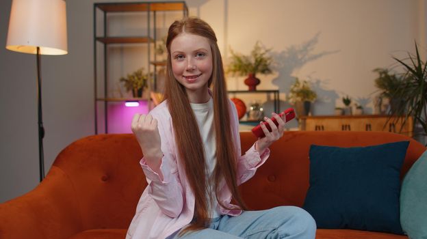 Happy excited joyful redhead teen child girl use mobile smartphone typing browsing say Wow yes found out great big lottery win, good news, celebrate sitting at home couch. Ginger female children kid