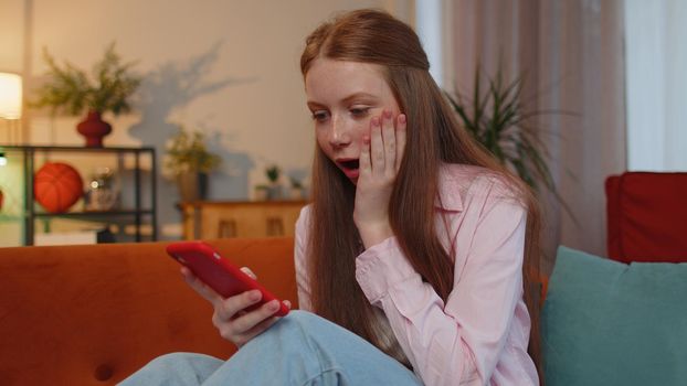 Oh my God, Wow. Happy excited teen young child girl use mobile smartphone typing browsing say yes found out great big jackpot lottery win, good news, celebrate at home couch. Ginger children kid alone