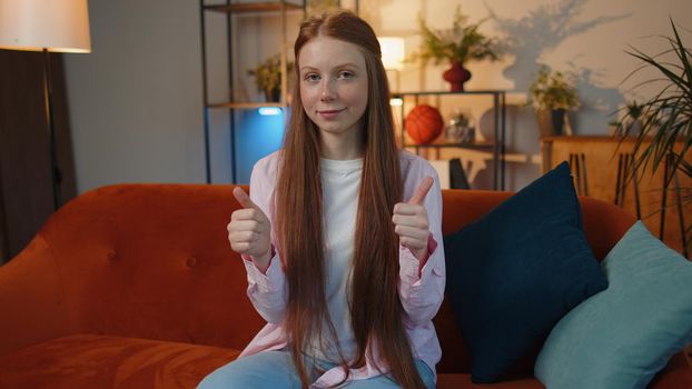 Like. Happy young redhead teenager child girl looking approvingly at camera showing thumbs up, like sign positive something good, positive feedback sits at home room couch. Ginger female children kid