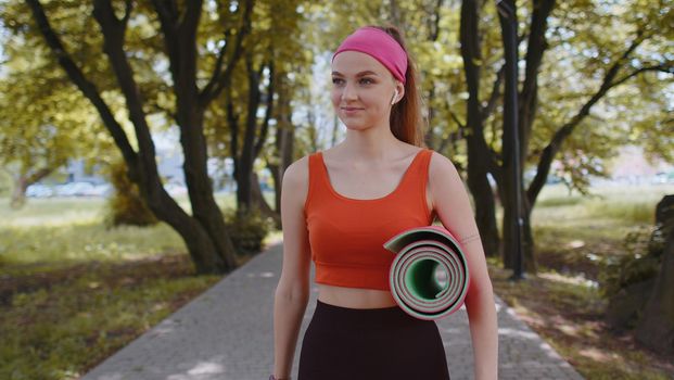 Athletic fitness sport runner girl walking with sport yoga mat to do fitness stretching exercises. Young woman training cardio workout in park at morning. Active sportswoman. Aerobic, weight loss