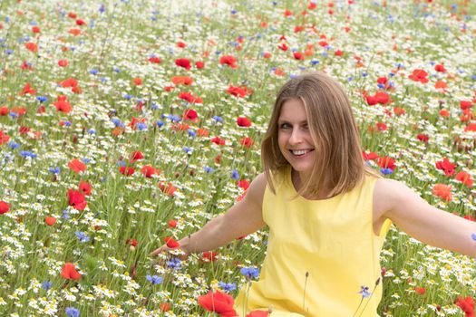 a beautiful young blonde woman in a yellow dress stands among a flowering field of poppies, daisies, cornflowers and laughs. High quality photo